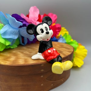 May include: A ceramic figurine of Mickey Mouse wearing red shorts with white polka dots. He is sitting on a wooden box with a colorful lei behind him.