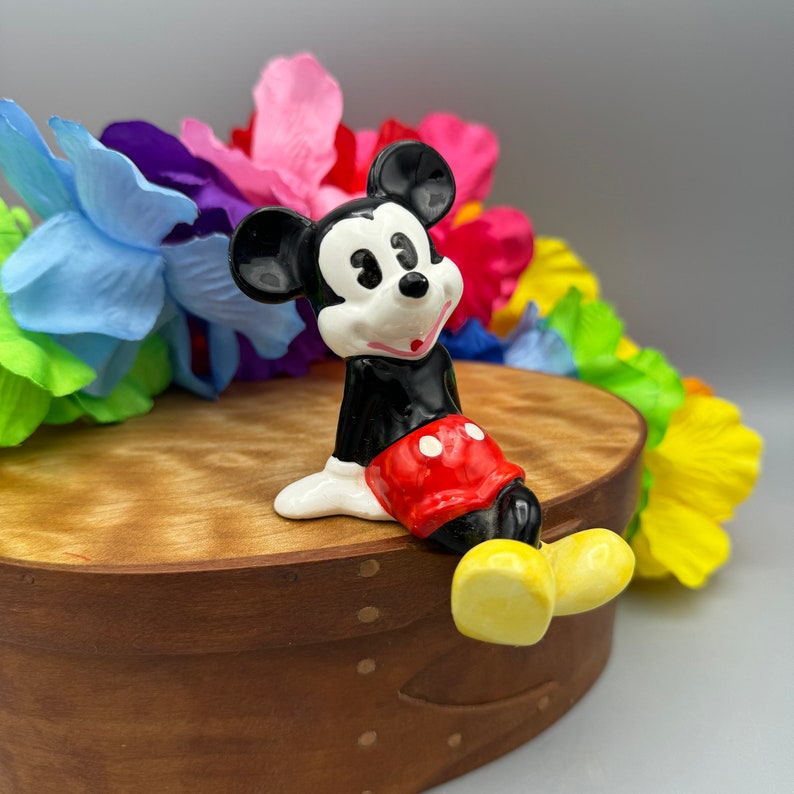 May include: A ceramic figurine of Mickey Mouse wearing red shorts with white polka dots. He is sitting on a wooden box with a colorful lei behind him.