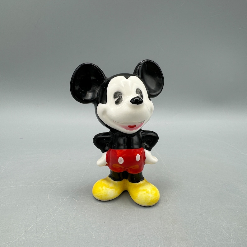 Może przedstawiać: Ceramiczna figurka Myszki Miki. Ma na sobie swoje charakterystyczne czerwone spodenki z czarnymi guzikami i ż&oacute;łte buty. Ma czarno-białą twarz z szerokim uśmiechem.