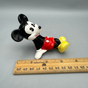 May include: A ceramic figurine of Mickey Mouse lying on his side. He is wearing his signature red shorts with white polka dots and yellow shoes. The figurine is approximately 2 inches long.