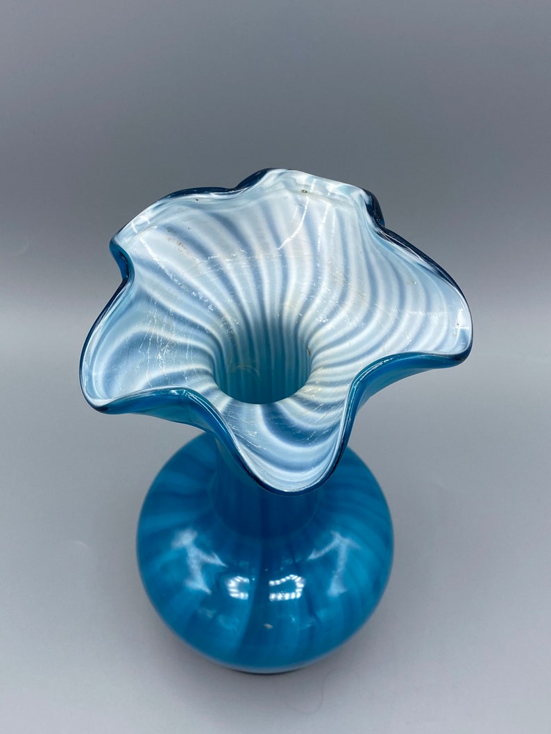 Puede incluir: Un jarr&oacute;n de vidrio azul con patrones de espiral blancos. El jarr&oacute;n tiene una abertura ancha y rizada y un cuerpo redondo.