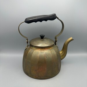 May include: A vintage brass kettle with a black plastic handle. The kettle has a rounded body with vertical ridges and a spout on the side.