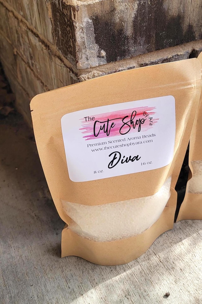 May include: A brown paper bag with a clear window, filled with white aroma beads. The bag has a label with the text "The Cute Shop" and "Diva". The label also includes the text "Premium Scented Aroma Beads" and "8 oz."