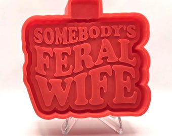 May include: A red cookie cutter with the words "SOMEBODY'S FERAL WIFE" in a bold, retro font. The cutter has a rectangular shape with a scalloped edge and a small handle at the top.