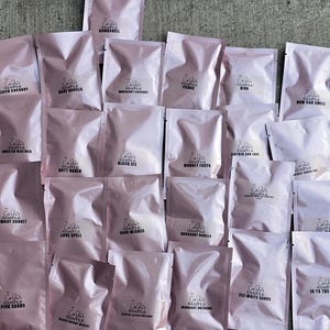 May include: A collection of pink sample packets, each labeled with a fragrance name in black text. The packets are arranged on a gray concrete surface. Fragrance names include "Black Coconut," "Sweater Weather," and "Love Spell."