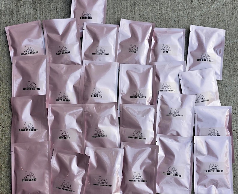 May include: A collection of pink sample packets, each labeled with a fragrance name in black text. The packets are arranged on a gray concrete surface. Fragrance names include "Black Coconut," "Sweater Weather," and "Love Spell."