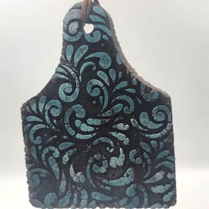 May include: A dark, rectangular leather tag with a teal floral pattern. The tag is suspended by a brown leather cord. The design is a repeating pattern of swirls and leaves.