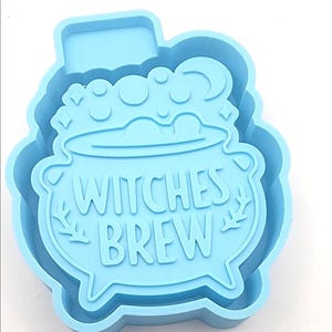 May include: A light blue silicone mold in the shape of a cauldron with the words "Witches Brew" in a whimsical font. The cauldron has bubbles and a crescent moon design. The mold is for crafting.