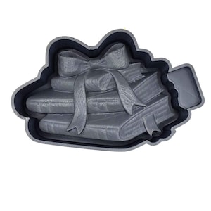 May include: A gray cookie cutter in the shape of a stack of books tied with a bow. The cutter has a scalloped edge and is made of plastic. Ideal for making book-themed cookies.