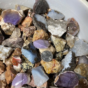 May include: A collection of colorful agate stones in various shapes and sizes. The stones are a mix of purple, blue, white, brown, and gray. A US quarter is included for scale.