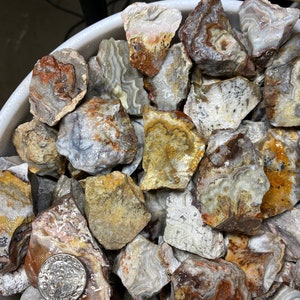May include: A white bowl filled with rough-cut agate stones. The stones display a variety of colors, including shades of gray, brown, orange, and white. The agate pieces have unique patterns and textures, with some showing banded formations.