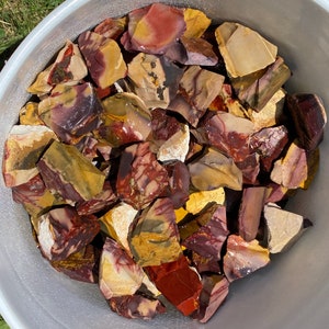 May include: A close-up shot of a white bucket filled with a variety of polished rocks. The rocks display a mix of colors, including red, yellow, brown, and purple. The stones have a smooth, glossy finish, suggesting they have been tumbled or polished.