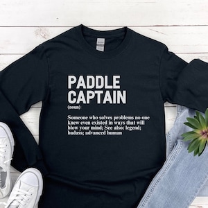 May include: Black long-sleeve t-shirt with white text that reads "PADDLE CAPTAIN (noun) Someone who solves problems no one knew even existed in ways that will blow your mind; See also: legend; badass; advanced human"