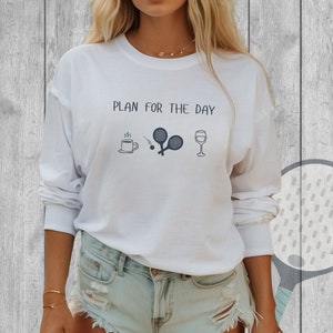 May include: White long-sleeve sweatshirt with the text "PLAN FOR THE DAY" printed on the front. The design includes a cup of coffee, two paddle tennis rackets, a ball, and a glass of wine.
