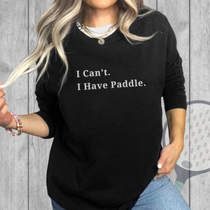 May include: Black long-sleeve sweatshirt with the text "I Can't. I Have Paddle." in white. The sweatshirt is worn by a person with blonde hair and a gold necklace. The background is a light gray wood panel.