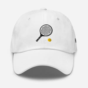May include: White baseball cap with an embroidered black and white paddle and yellow ball. The cap has a curved brim and an adjustable strap.