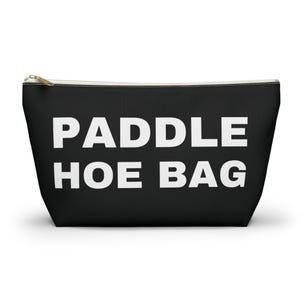 May include: Black zippered pouch with the text "PADDLE HOE BAG" in large white capital letters. The bag has a gold-colored zipper and a rectangular shape. The pouch is likely made of a durable fabric.