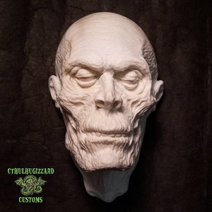 May include: A white plaster cast of a human head, sculpted to resemble a mummy. The head is tilted slightly to the right, and the eyes are closed. The mouth is slightly open, and the lips are parted. The head is detailed with wrinkles and lines, giving it a realistic appearance.