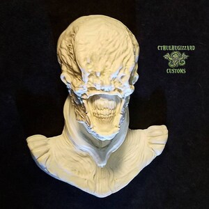 May include: A white, detailed, sculpted bust of a creature with a large, open mouth and a textured, wrinkled face. The bust is on a black background. The text "CTHULHUGIZZARD CUSTOMS" is visible in the upper right corner of the image.