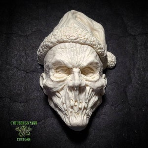 May include: A white ceramic sculpture of a face wearing a Santa hat. The face has a grotesque expression with a wide open mouth and sharp teeth. The sculpture is detailed with wrinkles and folds in the skin.