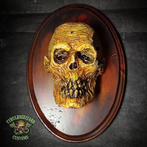 May include: A yellow, detailed, sculpted human skull with a toothy grin, mounted on a brown oval wooden plaque. The plaque has a dark brown stain in the center and a logo at the bottom that reads "Cthulhuizzard Customs".