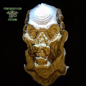 K&ouml;nnte beinhalten: Eine detaillierte, handbemalte, wei&szlig;e Gips-Skulptur eines monstr&ouml;sen, skelettartigen Gesichts mit einem aufgerissenen Mund und einer strukturierten, faltigen Oberfl&auml;che. Die Skulptur wird von oben beleuchtet, wodurch die filigranen Details hervorgehoben werden.
