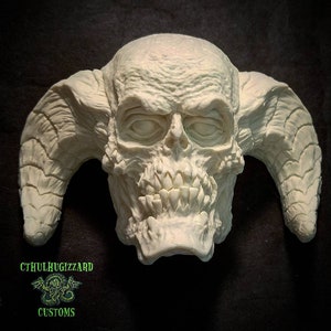May include: A white resin sculpture of a demonic skull with horns. The skull has a detailed, textured surface and a menacing expression. The horns are curved and pointed.