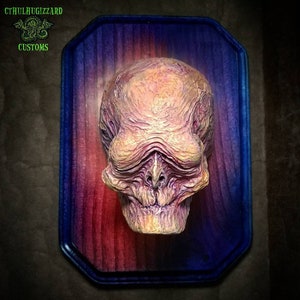 May include: A grotesque, textured sculpture of a humanoid head with a prominent brow and a skeletal grin. The sculpture is mounted on a wooden plaque with a blue border and a red and purple gradient. The text "CTHULAUGIZZARD CUSTOMS" is in the upper left corner.