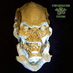 K&ouml;nnte beinhalten: Eine detaillierte, handbemalte, beige und wei&szlig;e Zombie-Maske mit einem knurrenden Ausdruck. Die Maske ist mit realistischen Details wie Falten, Z&auml;hnen und einer strukturierten Oberfl&auml;che modelliert. Die Maske tr&auml;gt den Titel "CthulhuGizzard Customs".