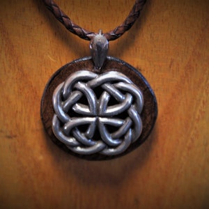 Könnte beinhalten: Ein silberner keltischer Knotenanhänger an einer braunen Lederkordel. Der Anhänger ist rund und hat eine Holzrückseite.