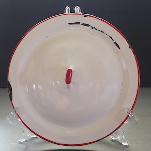 May include: A white enamelware plate with a red rim and handle. The plate has some black paint chipping around the rim. The handle is a small, red, rounded shape. The plate is displayed on a clear plastic stand.