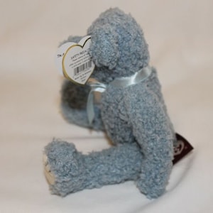 May include: A light blue plush teddy bear with a blue ribbon around its neck. The bear is sitting on a white surface. The bear has a safety tag attached to its ear.