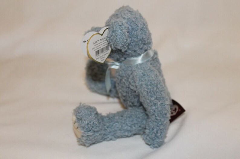 May include: A light blue plush teddy bear with a blue ribbon around its neck. The bear is sitting on a white surface. The bear has a safety tag attached to its ear.