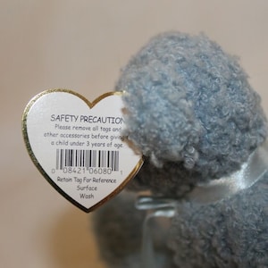 May include: A heart-shaped tag with a safety precaution message and a barcode. The tag is attached to a gray plush toy. The tag reads "SAFETY PRECAUTION Please remove all tags and other accessories before giving a child under 3 years of age. 0 08421 06080 1 Retain Tag For Reference Surface Wash"
