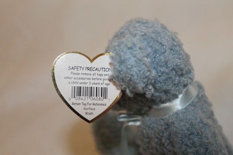 May include: A heart-shaped tag with a safety precaution message and a barcode. The tag is attached to a gray plush toy. The tag reads "SAFETY PRECAUTION Please remove all tags and other accessories before giving a child under 3 years of age. 0 08421 06080 1 Retain Tag For Reference Surface Wash"