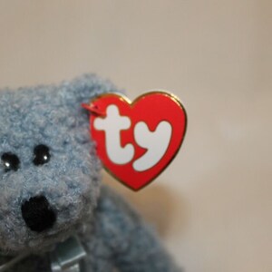 May include: A blue plush teddy bear with a Ty heart-shaped tag attached to its ear. The tag is red with a white "ty" logo.