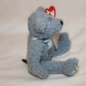 May include: A light blue plush teddy bear with a blue ribbon around its neck. The bear has a red heart-shaped tag attached to its ear.