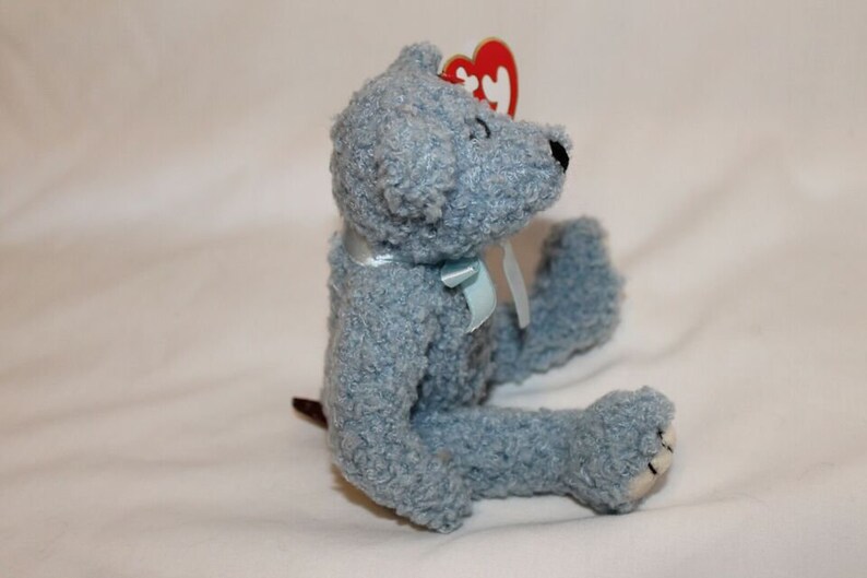 May include: A light blue plush teddy bear with a blue ribbon around its neck. The bear has a red heart-shaped tag attached to its ear.