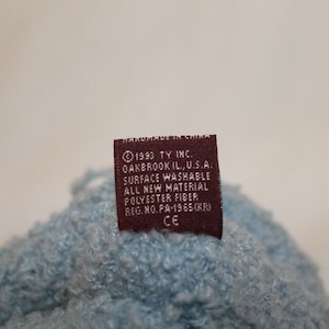 May include: A brown label with white text that reads "1993 TY INC. OAKBROOK IL., U.S.A. SURFACE WASHABLE ALL NEW MATERIAL POLYESTER FIBER REG. NO. PA-1965(KR) CE" is attached to a light blue fuzzy fabric.