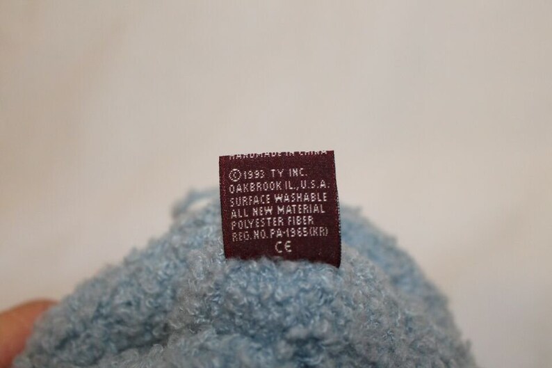 May include: A brown label with white text that reads "1993 TY INC. OAKBROOK IL., U.S.A. SURFACE WASHABLE ALL NEW MATERIAL POLYESTER FIBER REG. NO. PA-1965(KR) CE" is attached to a light blue fuzzy fabric.