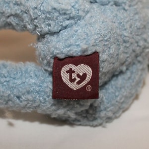 May include: A light blue plush toy with a Ty heart-shaped tag attached. The tag is brown with white stitching and the Ty logo in white.