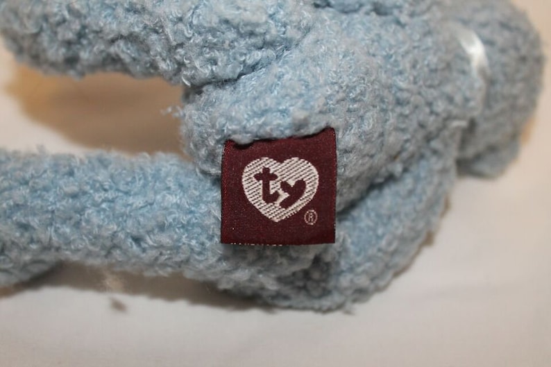 May include: A light blue plush toy with a Ty heart-shaped tag attached. The tag is brown with white stitching and the Ty logo in white.