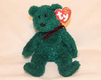 May include: Green plush beanie baby bear with a red velvet ribbon around its neck. The bear has black eyes and a black nose. The bear is sitting on a white surface. The bear has a Ty tag attached to its ear.