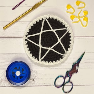 Pode incluir: Um descanso de copo de crochê preto e branco com um design de estrela de cinco pontas. O descanso de copo é rodeado por marcadores de ponto amarelos, um gancho de crochê dourado, um gancho de crochê roxo, uma fita métrica azul e tesouras de unicórnio arco-íris.