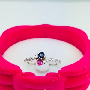 May include: A silver ring with a blue and red gemstone setting. The ring is set in a white velvet box.