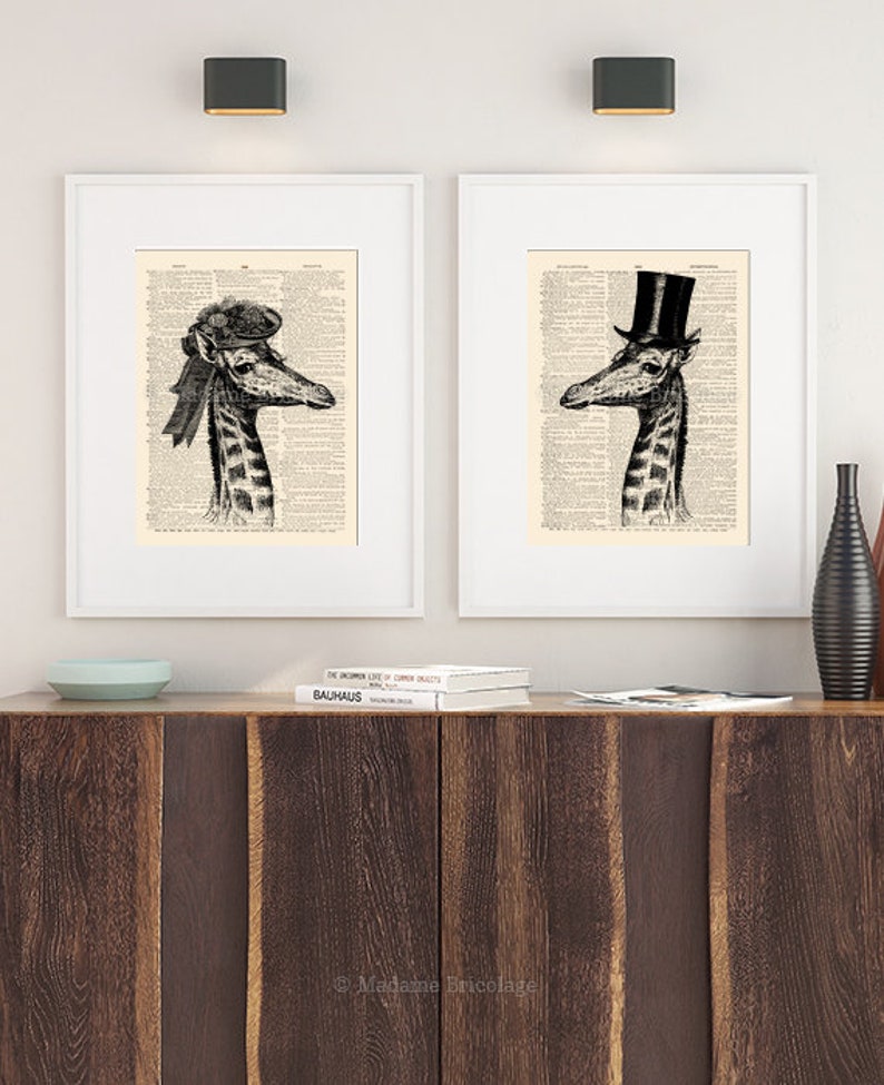 May include: Two framed black and white illustrations of giraffes wearing hats. The giraffe on the left is wearing a black and white hat with a ribbon. The giraffe on the right is wearing a black top hat. Both illustrations are printed on a vintage dictionary page background.