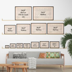 May include: A white wall with a shelf displaying various sizes of framed black and white prints. The prints are all of the same design, a vintage newspaper article. The sizes are listed in inches and centimeters. The shelf is made of wood and is mounted on the wall. There is a gray couch with pillows in front of the shelf. The couch is in a living room setting with a green plant and a coffee table.