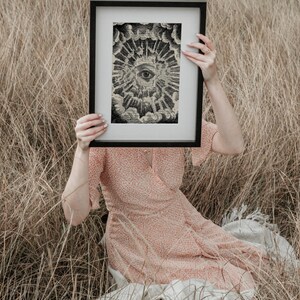 May include: A framed black and white print of an eye surrounded by clouds and rays of light. The print is held in front of a person wearing a pink floral dress.
