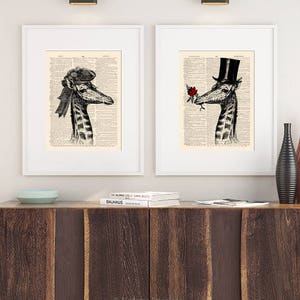 May include: Two framed prints of a giraffe wearing a hat and holding a rose. The prints are on a white background and are hanging on a wall. The wall is white and there is a wooden shelf below the prints.