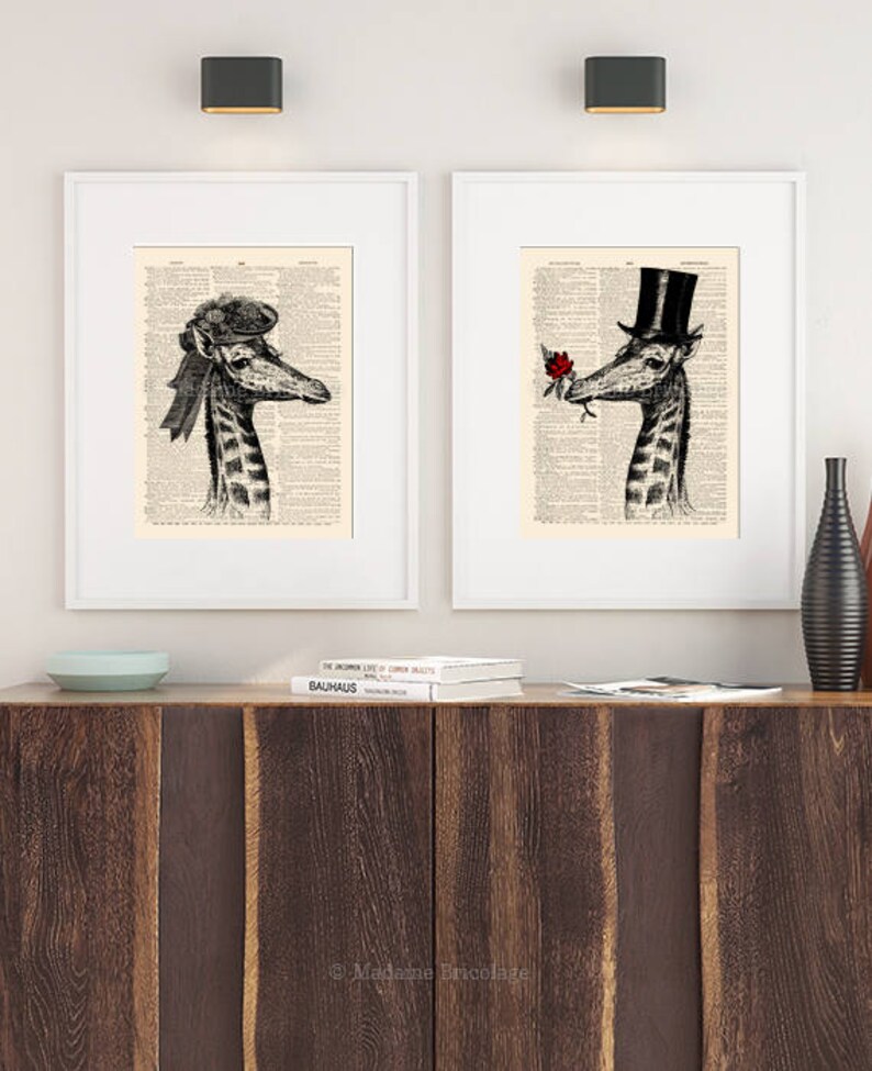 May include: Two framed prints of a giraffe wearing a hat and holding a rose. The prints are on a white background and are hanging on a wall. The wall is white and there is a wooden shelf below the prints.